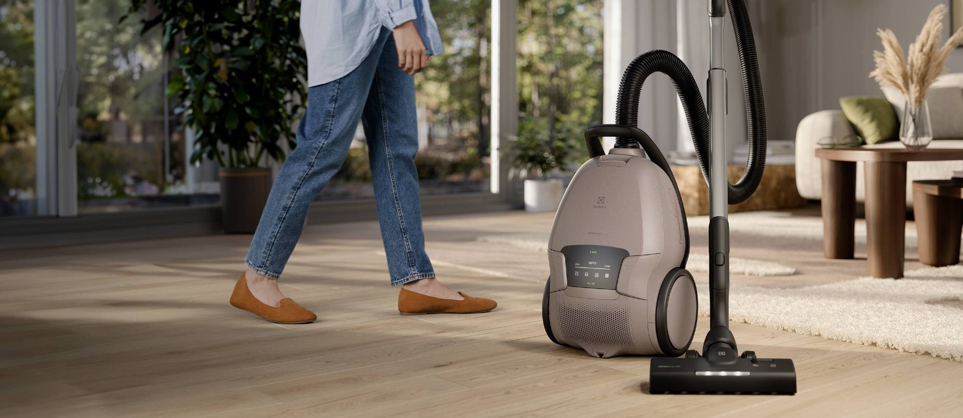 En støvsuger står midt på et stuegulv. En dame går forbi i bakgrunnen.